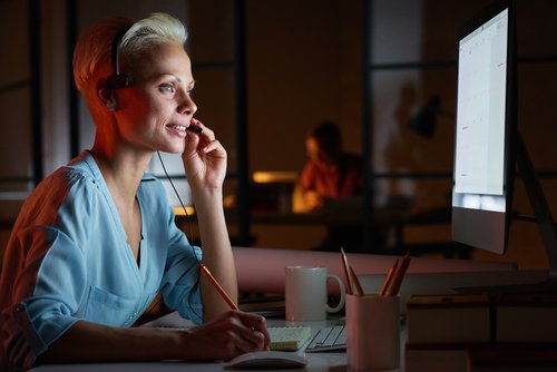 Una mujer joven con auriculares sentada en su lugar de trabajo y trabajando con el ordenador Young woman in headphones working on computer while sitting at her workplace