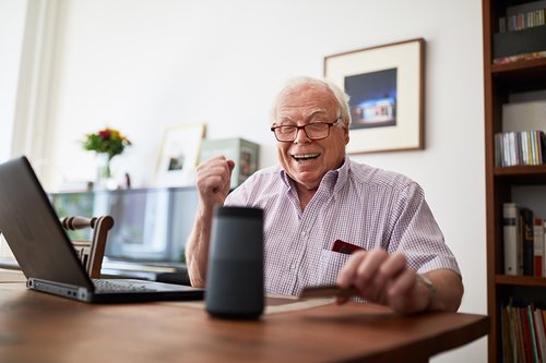 Retired man purchasing online using wireless devices  Senior man doing online shopping using a credit card, laptop and smart speaker device.